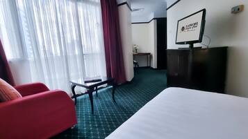 Quarto de hotel com uma cama, uma poltrona vermelha, uma mesinha, uma televisão sobre um suporte e uma janela com cortinas.