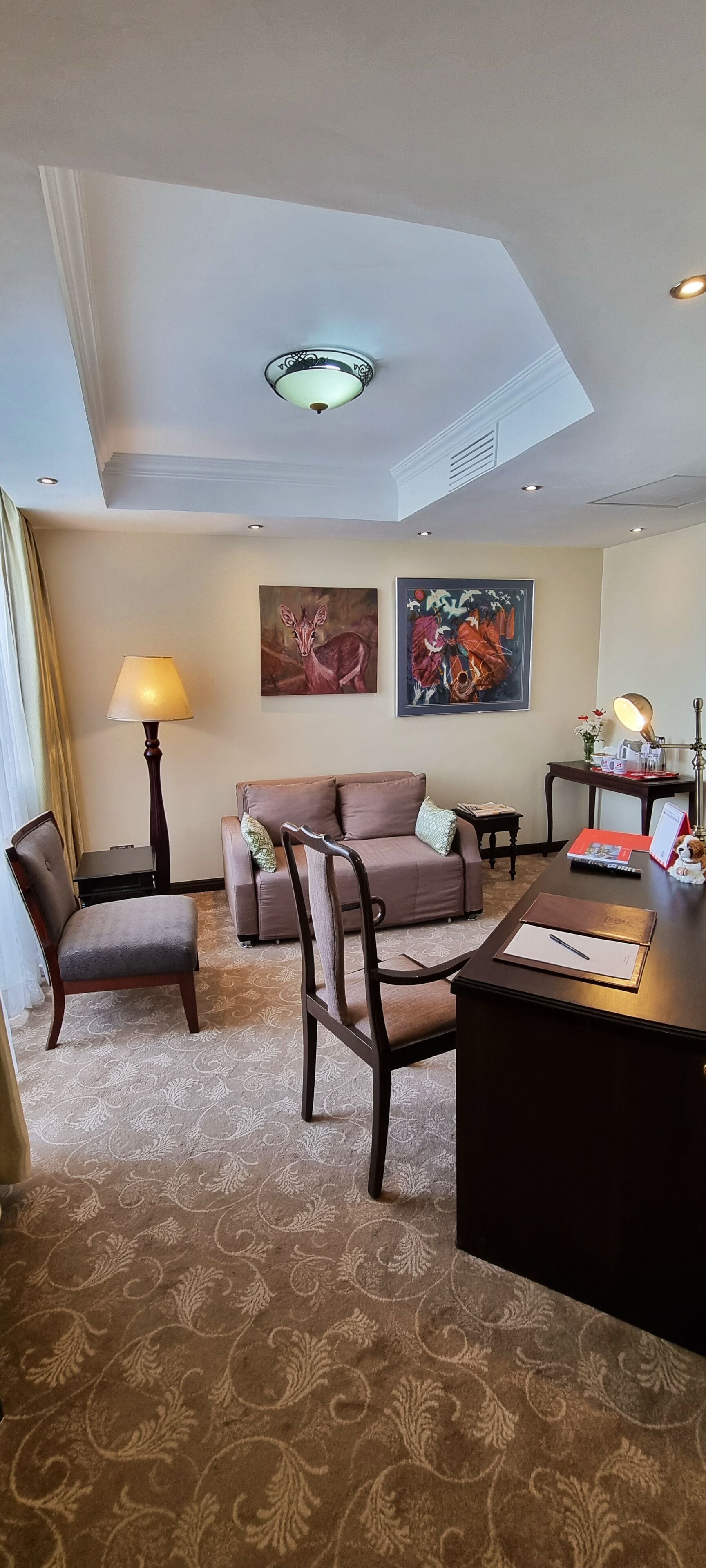 Habitación de hotel con alfombra estampada, un escritorio con lámpara, un sofá, dos sillones y obras de arte en las paredes.