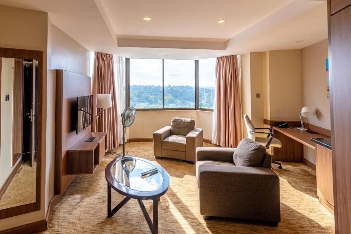A modern hotel room with a large window, a flat-screen TV, a sofa, a desk, and a fan.