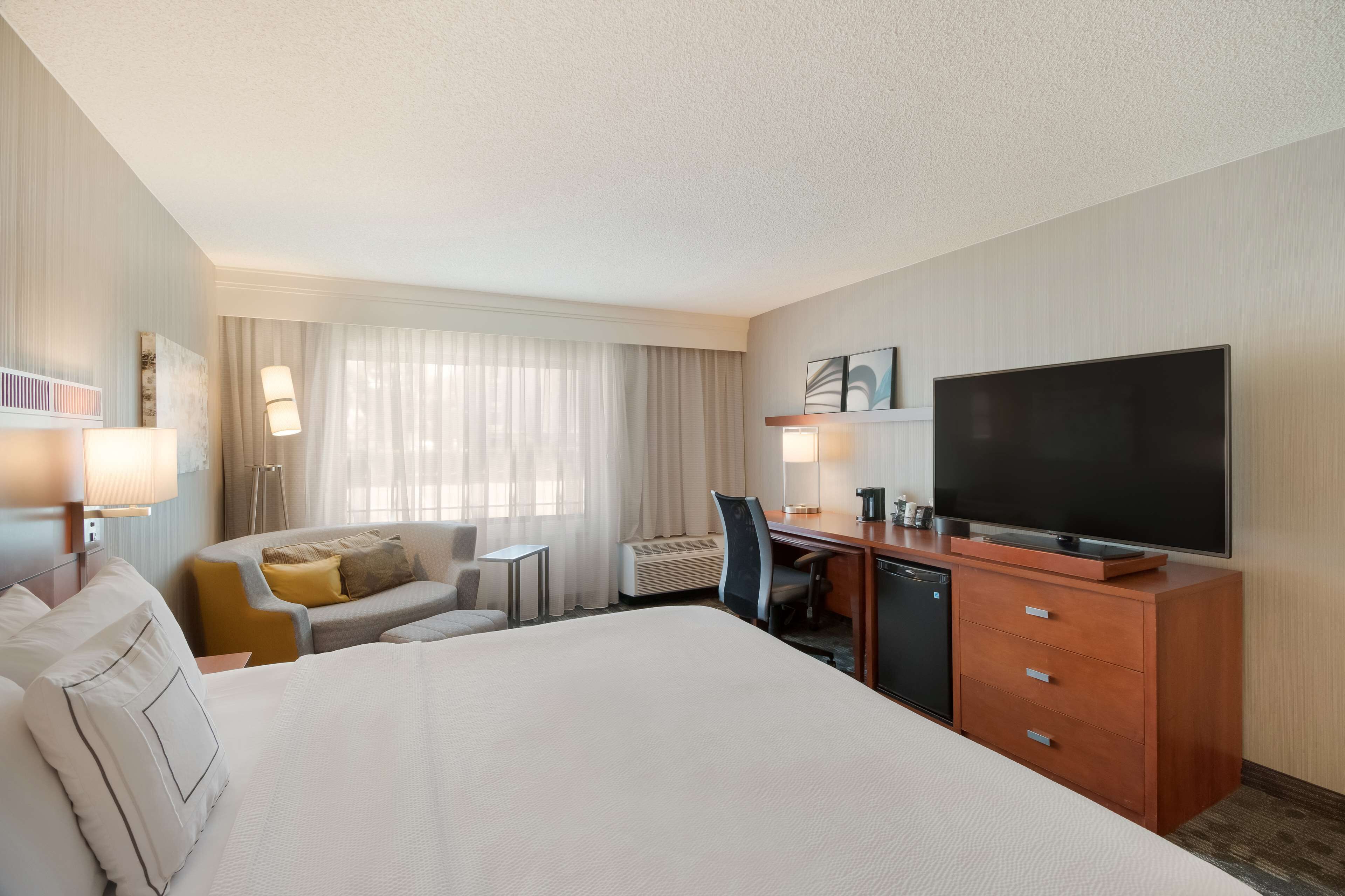 A hotel room with a large bed, a desk with a television, a sofa, and a window with curtains.