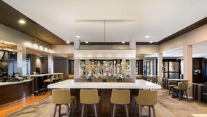 Un moderno vestíbulo de hotel con una zona de bar central, arreglos de asientos y un elemento decorativo.