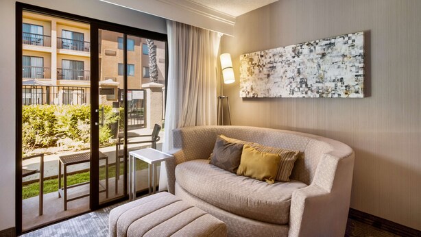 Habitación de hotel con sofá, reposapiés y una mesa. Se observa un ventanal con vistas a otros edificios y un balcón.