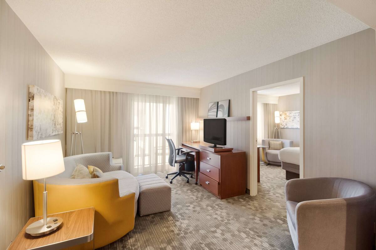 A hotel room with a sofa, a desk with a TV, a chair, and a lamp.