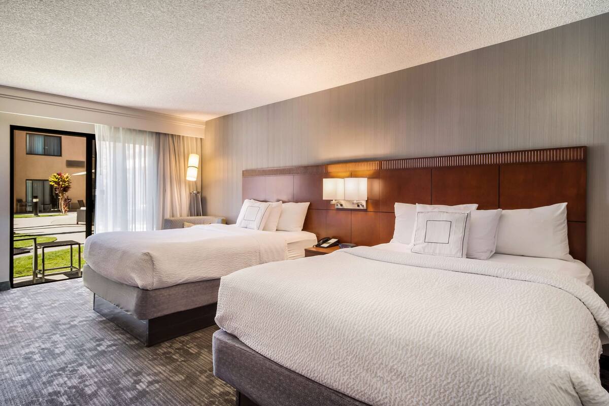 A hotel room with two beds, a large window with curtains, a view of the outdoors, and a small table with a lamp.