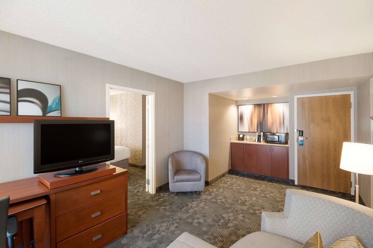 A hotel room with a flat-screen TV on a wooden dresser, a grey armchair, a sofa, a dining area with a microwave, and a wooden door.