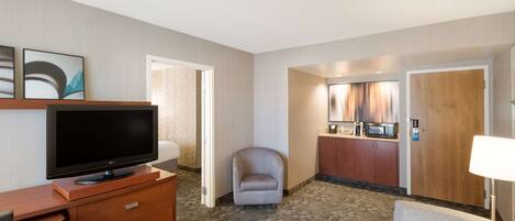 Habitación de hotel con televisión de pantalla plana sobre un tocador de madera, un sillón gris, un sofá, un rincón de comedor con microondas y una puerta de madera.