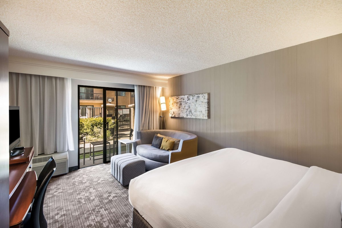 A hotel room with a large bed, a desk with a chair, a sofa, a small table, and a balcony with a view.
