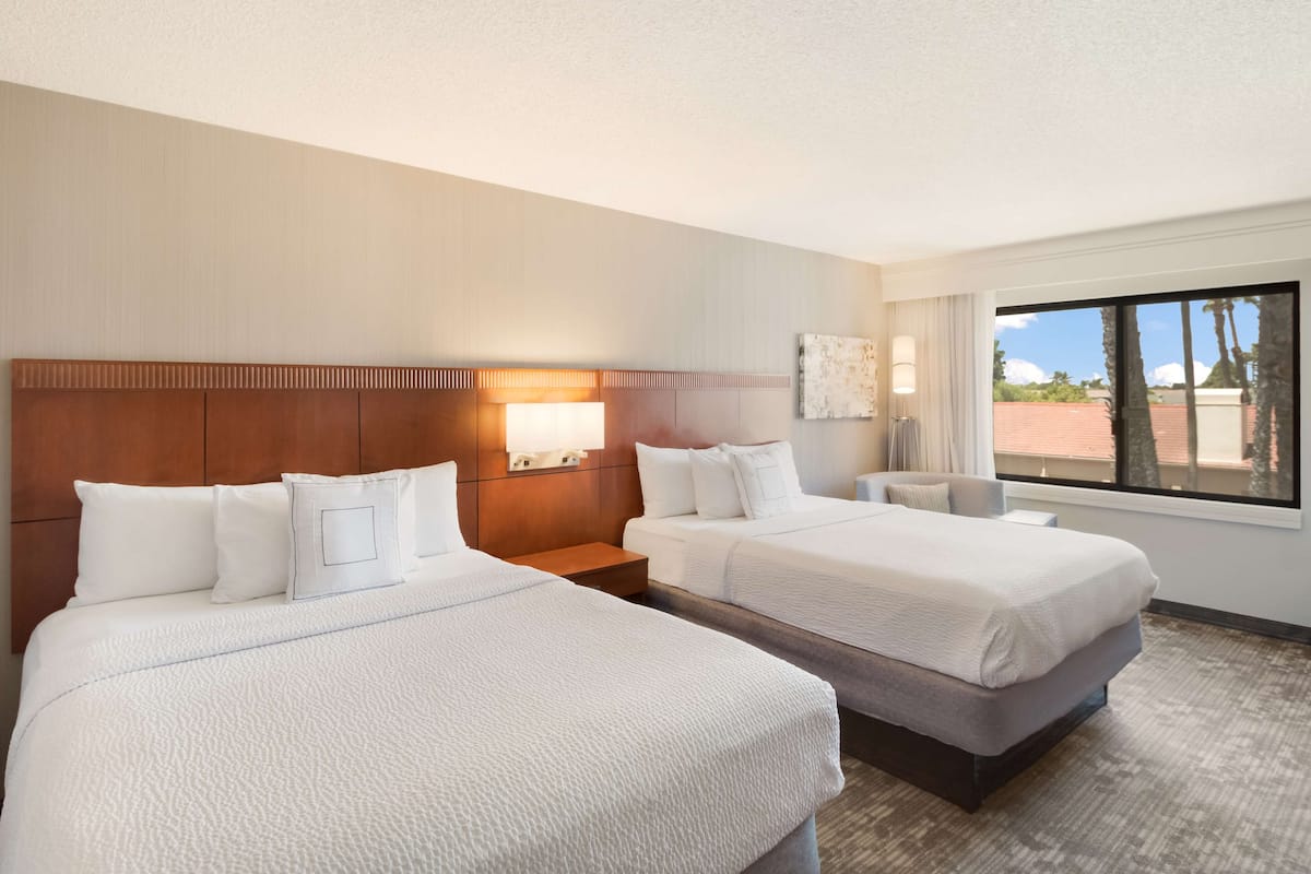A hotel room with two beds, a nightstand, a lamp, and a window with a view of palm trees.