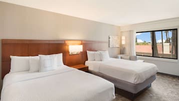 A hotel room with two beds, a nightstand, a lamp, and a window with a view of palm trees.