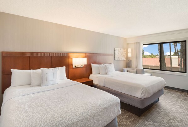 A hotel room with two beds, a nightstand, a lamp, and a window with a view of palm trees.