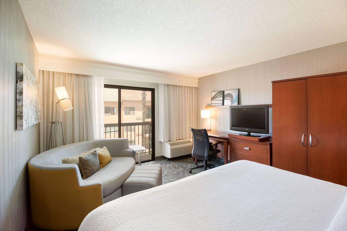 A hotel room with a bed, a sofa, a desk with a TV, and a balcony with curtains.