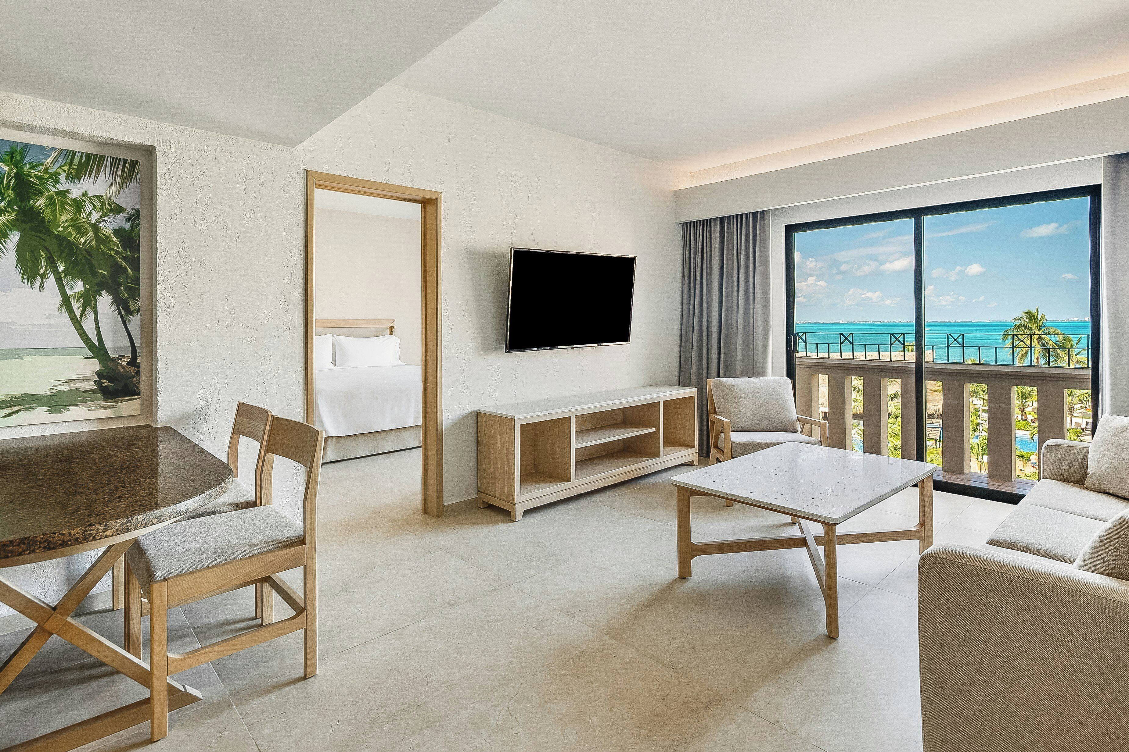 Quarto de hotel com televisão, mesa de jantar, cadeiras, sofá e varanda com vista para o oceano.