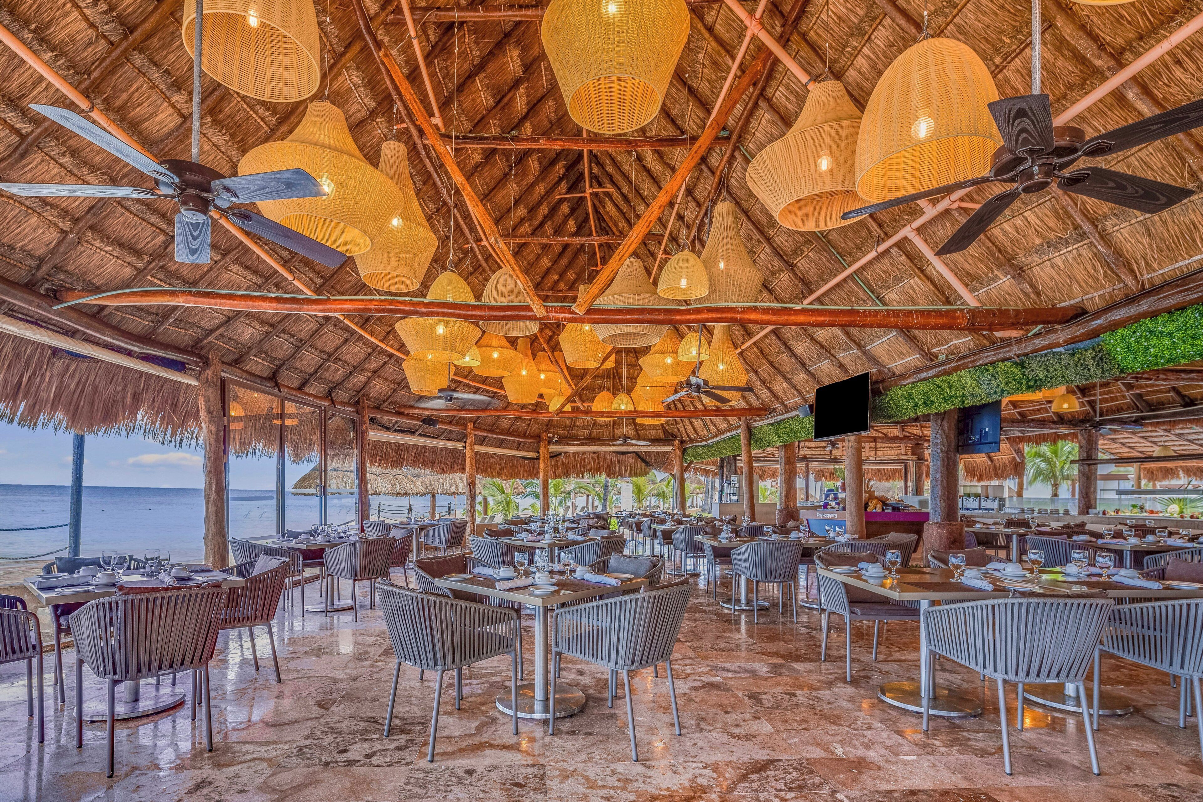A restaurant with thatched roofs, multiple dining tables, chairs, ceiling fans, and hanging lights.