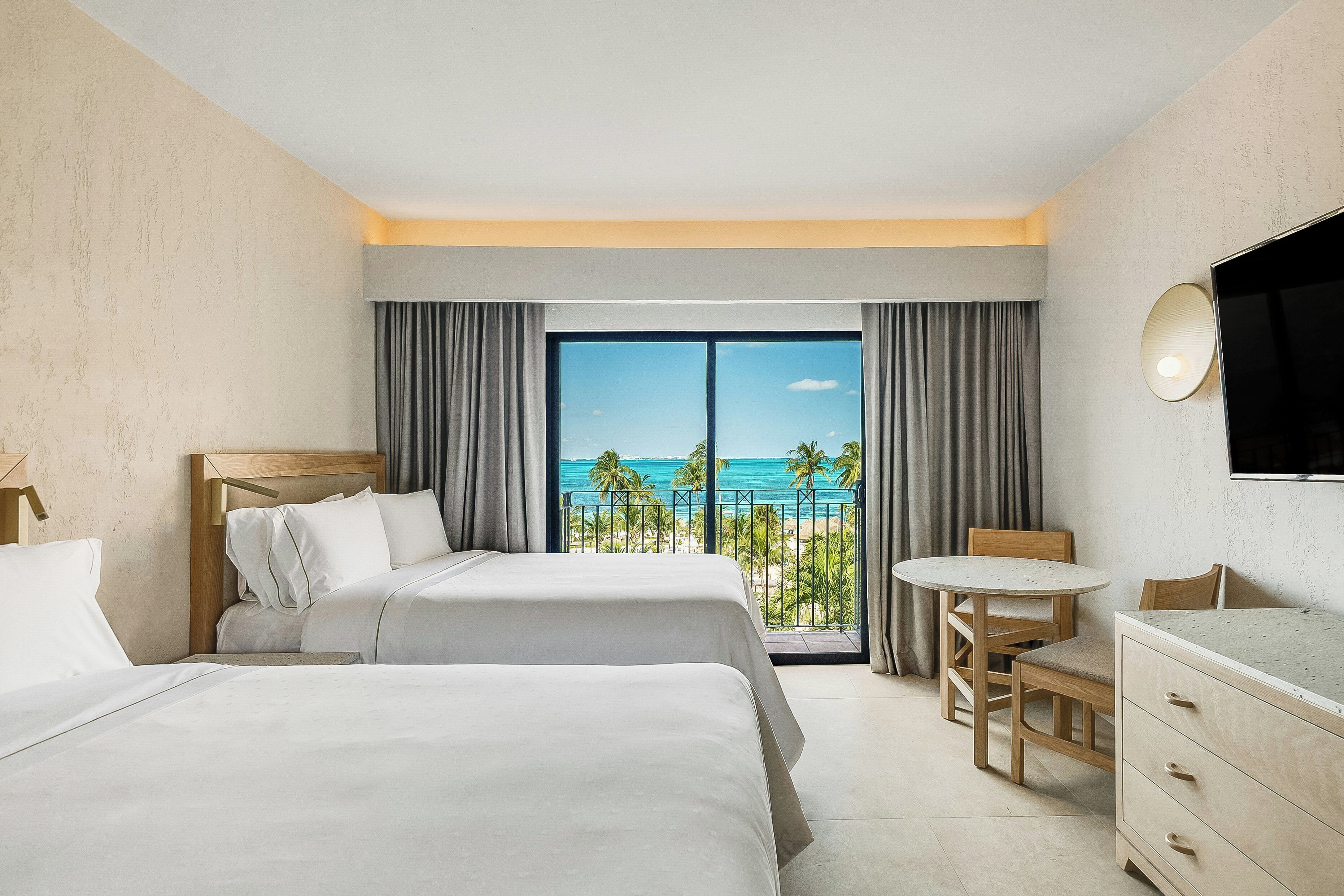 Ein Hotelzimmer mit zwei Betten, einem Schreibtisch, einem Sessel, einem Fernseher und einem Balkon mit Meerblick.