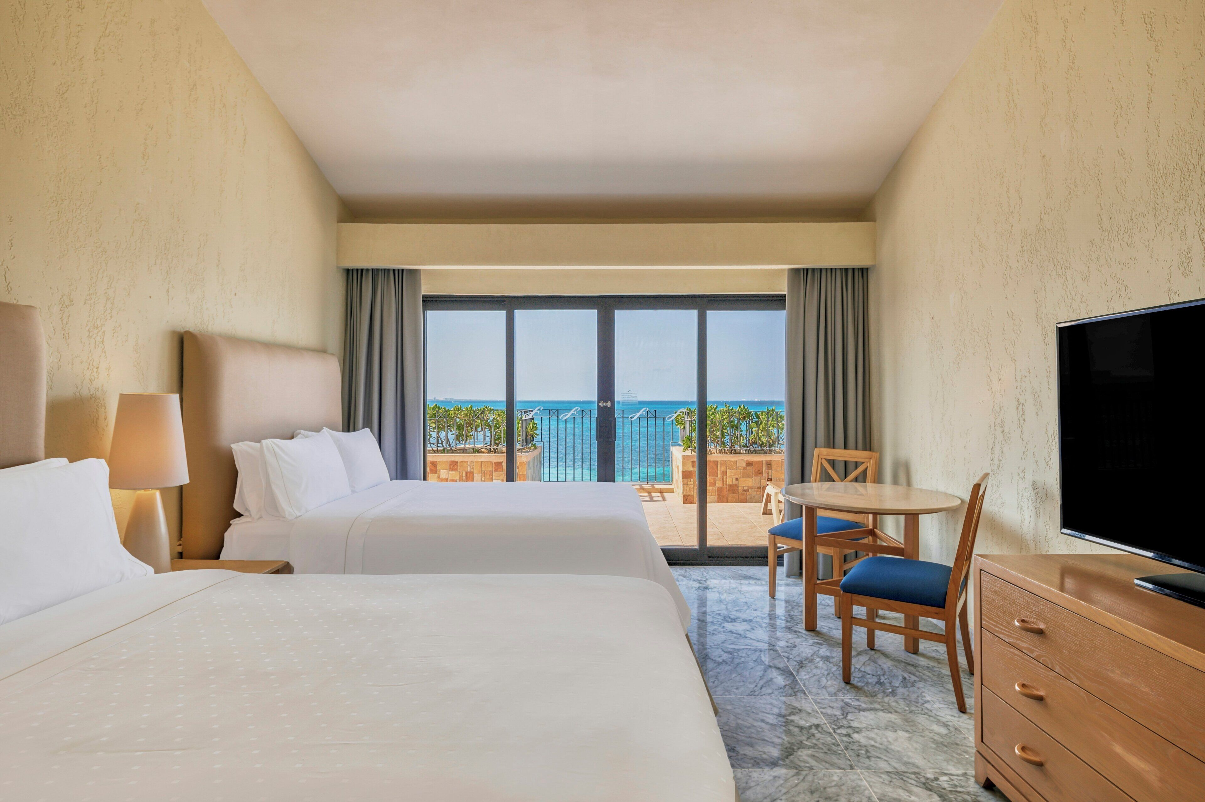 A hotel room with two beds, a TV, a desk with a chair, and a balcony with a view of the sea.