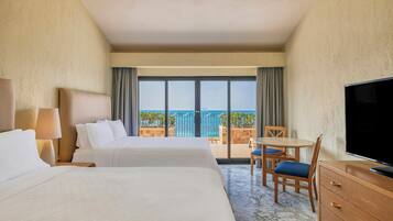 A hotel room with two beds, a TV, a desk with a chair, and a balcony with a view of the sea.