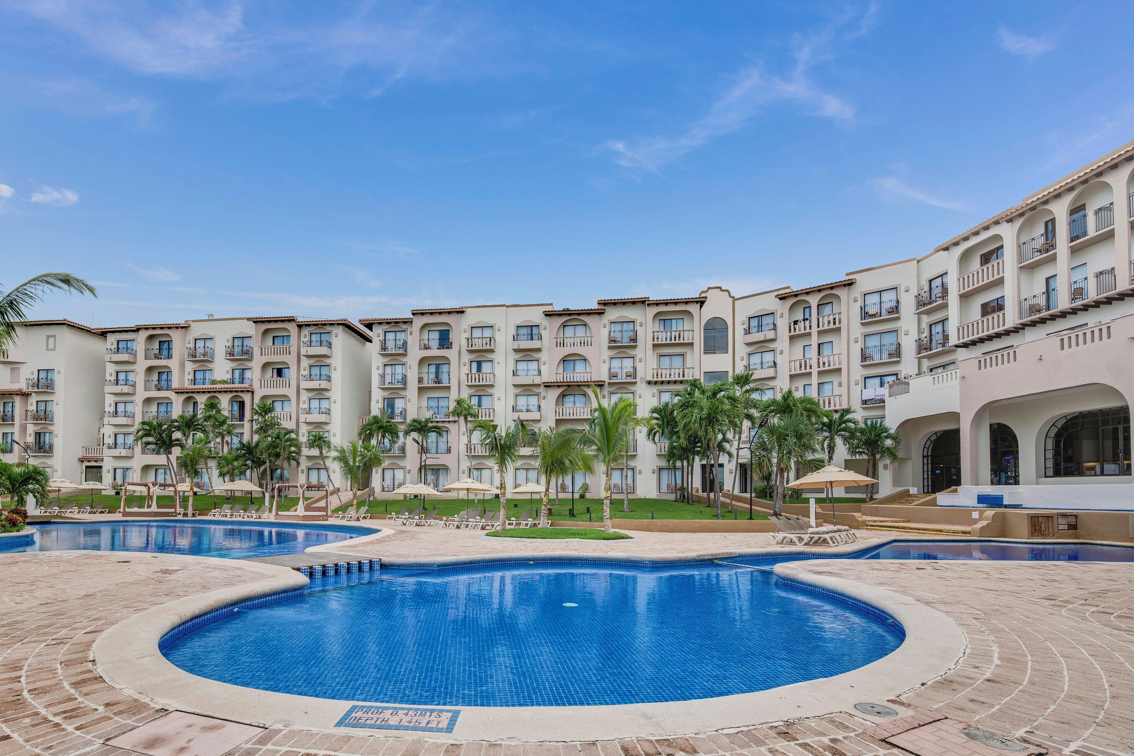 A hotel with multiple balconies, a central swimming pool, and palm trees.
