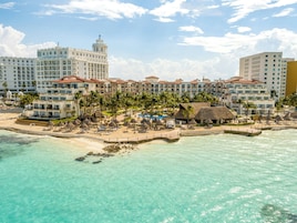Hotel frente al mar con varias edificaciones, un área de piscina y playa con sombrillas.