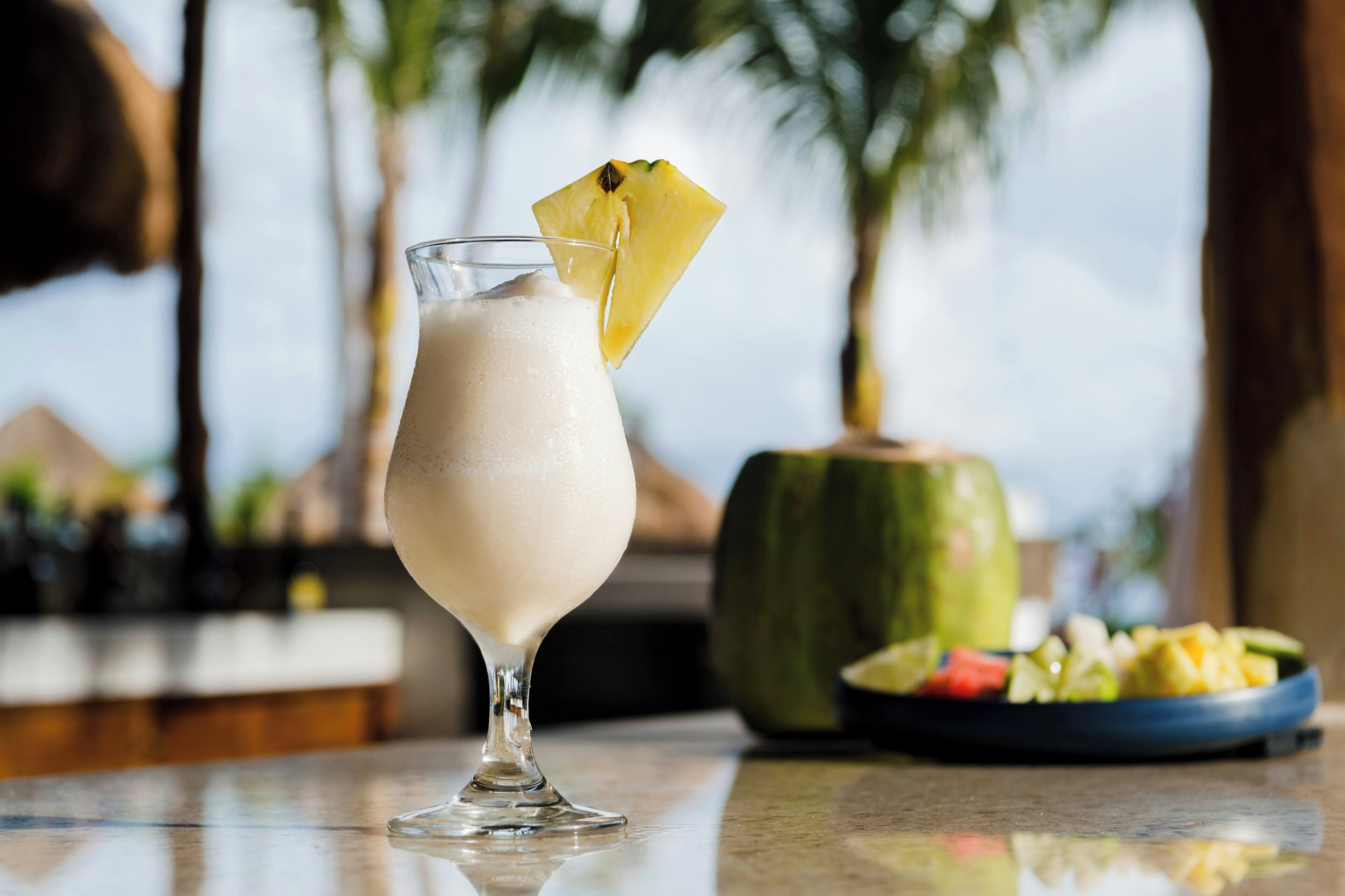 Un vaso de una bebida cremosa con piña como adorno, un plato de fruta y un recipiente verde, probablemente un coco, sobre una barra con un fondo desenfocado de palmeras.