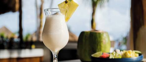 Un vaso de una bebida cremosa con piña como adorno, un plato de fruta y un recipiente verde, probablemente un coco, sobre una barra con un fondo desenfocado de palmeras.