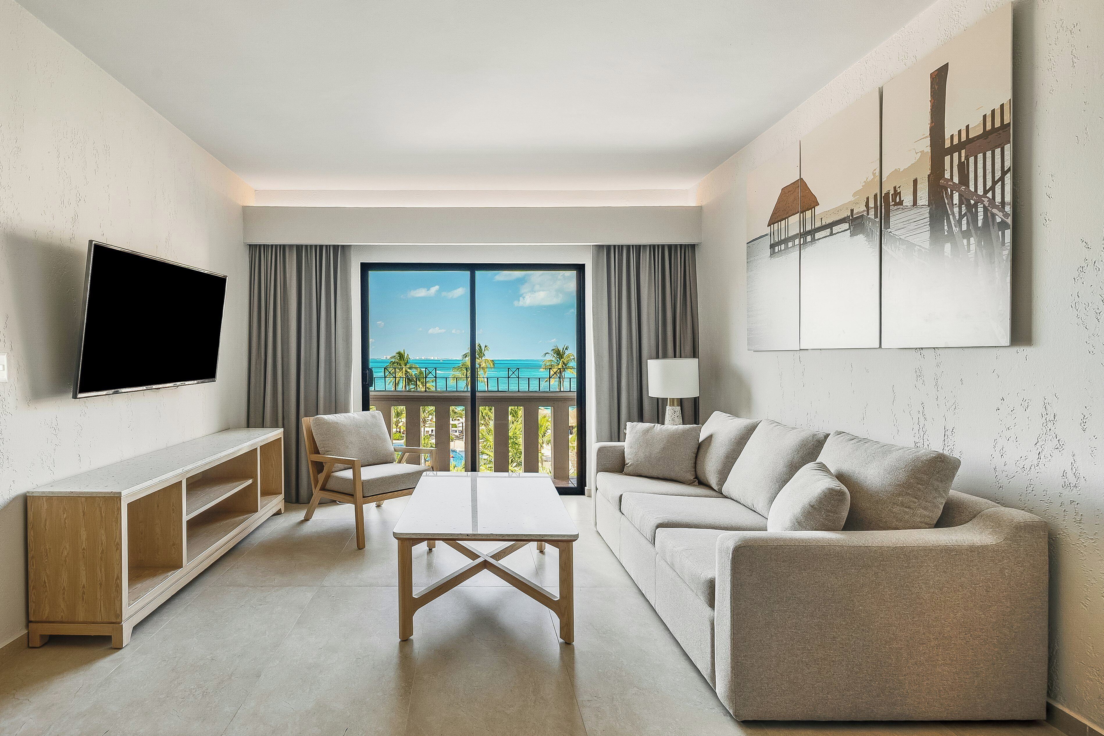 Ein modernes Wohnzimmer mit Flachbildfernseher, einer Couch, einem Couchtisch und Blick auf einen Strand und Palmen durch ein großes Fenster.