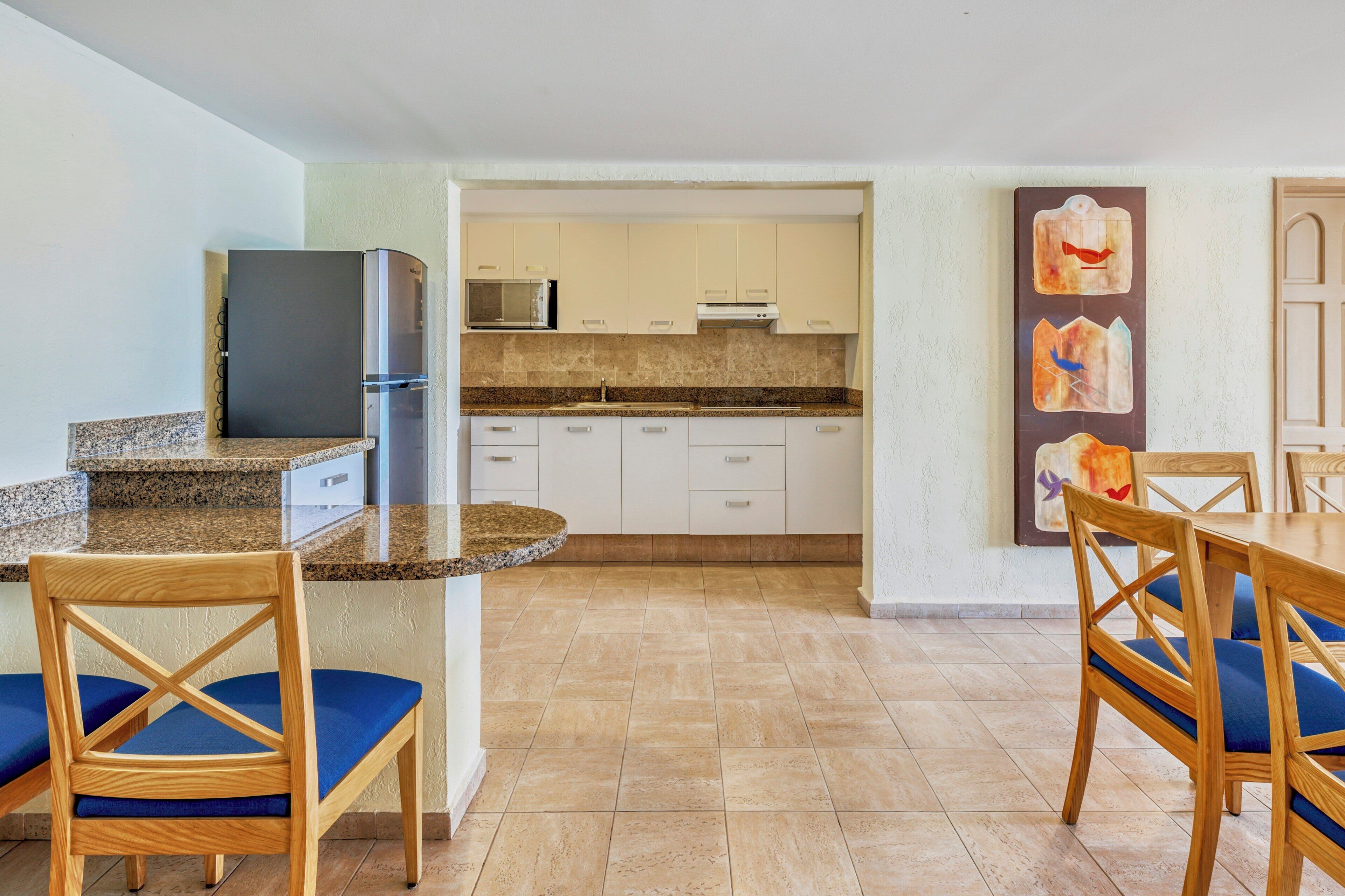 A kitchen with a refrigerator, microwave, and cabinets, a dining area with chairs, and a wall with artwork.