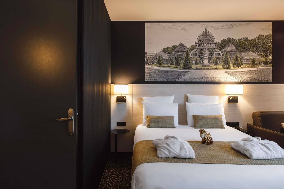 A hotel room with a large bed, two bedside lamps, a painting of a building, and a giraffe plush toy.