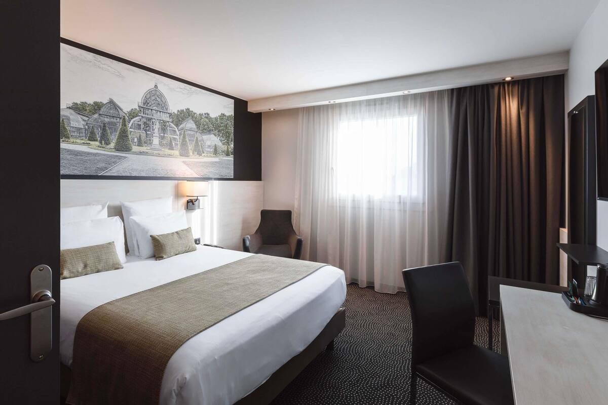A hotel room with a bed, a desk, a chair, a large window with curtains, and a wall-mounted picture.