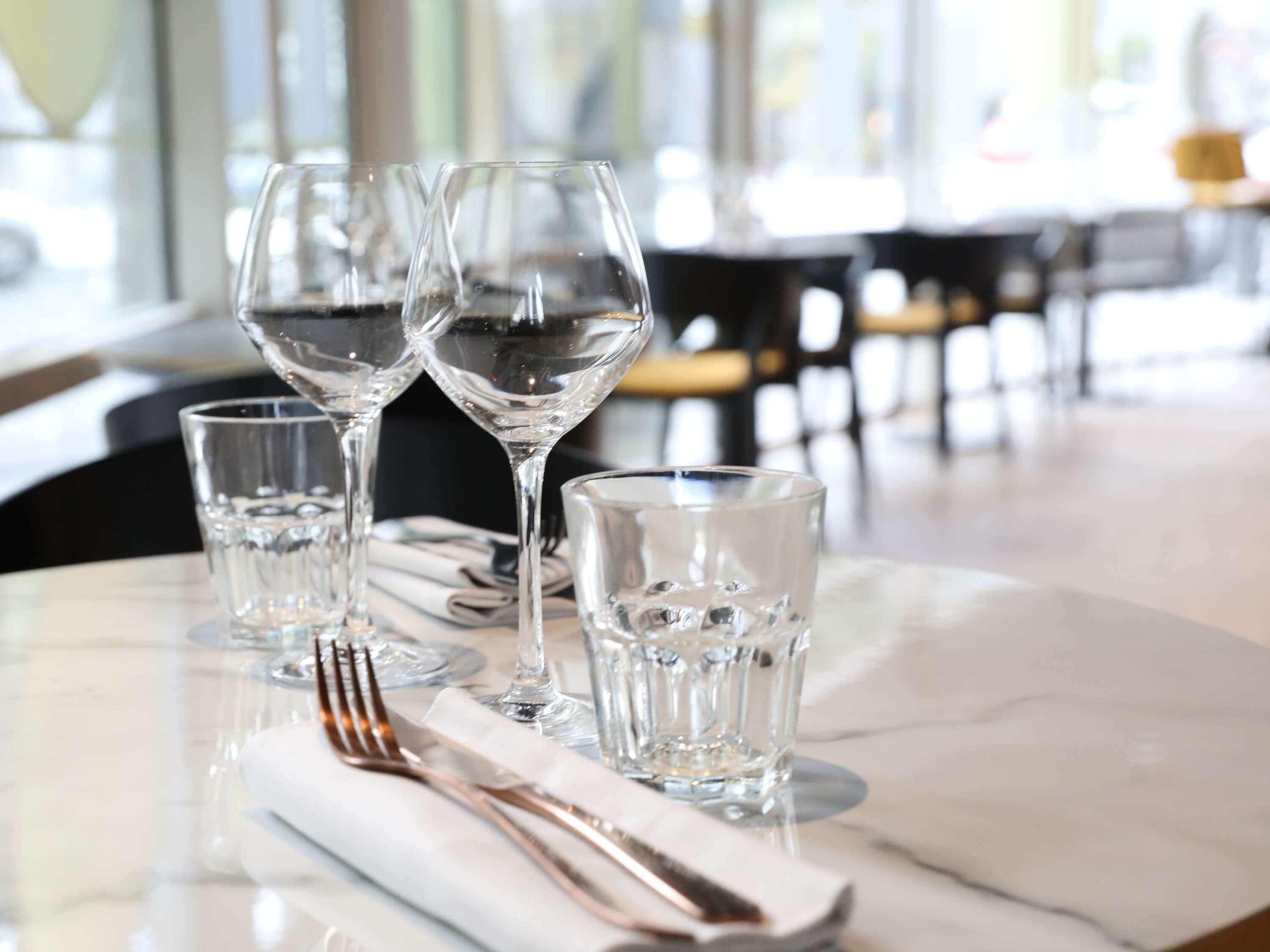 Une table dressée avec quatre verres à vin, un petit verre et des couverts, le tout posé sur une surface en marbre, dans un coin repas où l’on aperçoit des chaises et des tables en arrière-plan.