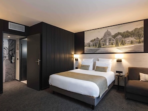 Une chambre d’hôtel avec un grand lit, un bureau avec une chaise, un téléviseur fixé au mur et une porte donnant sur un couloir.