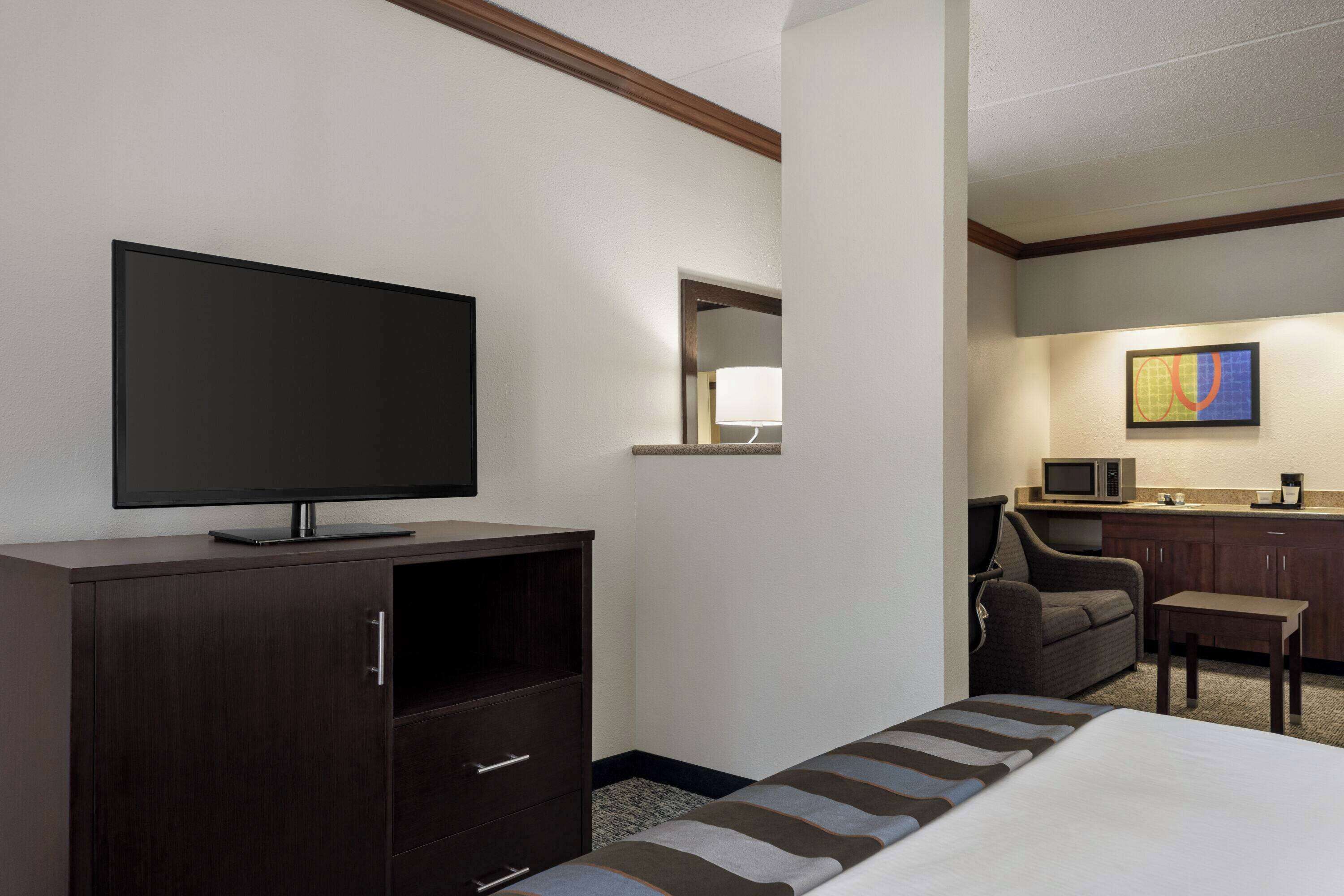 Una habitación de hotel con una cama, un televisor sobre un mueble de madera, una mesita, una silla, un microondas y una cafetera.