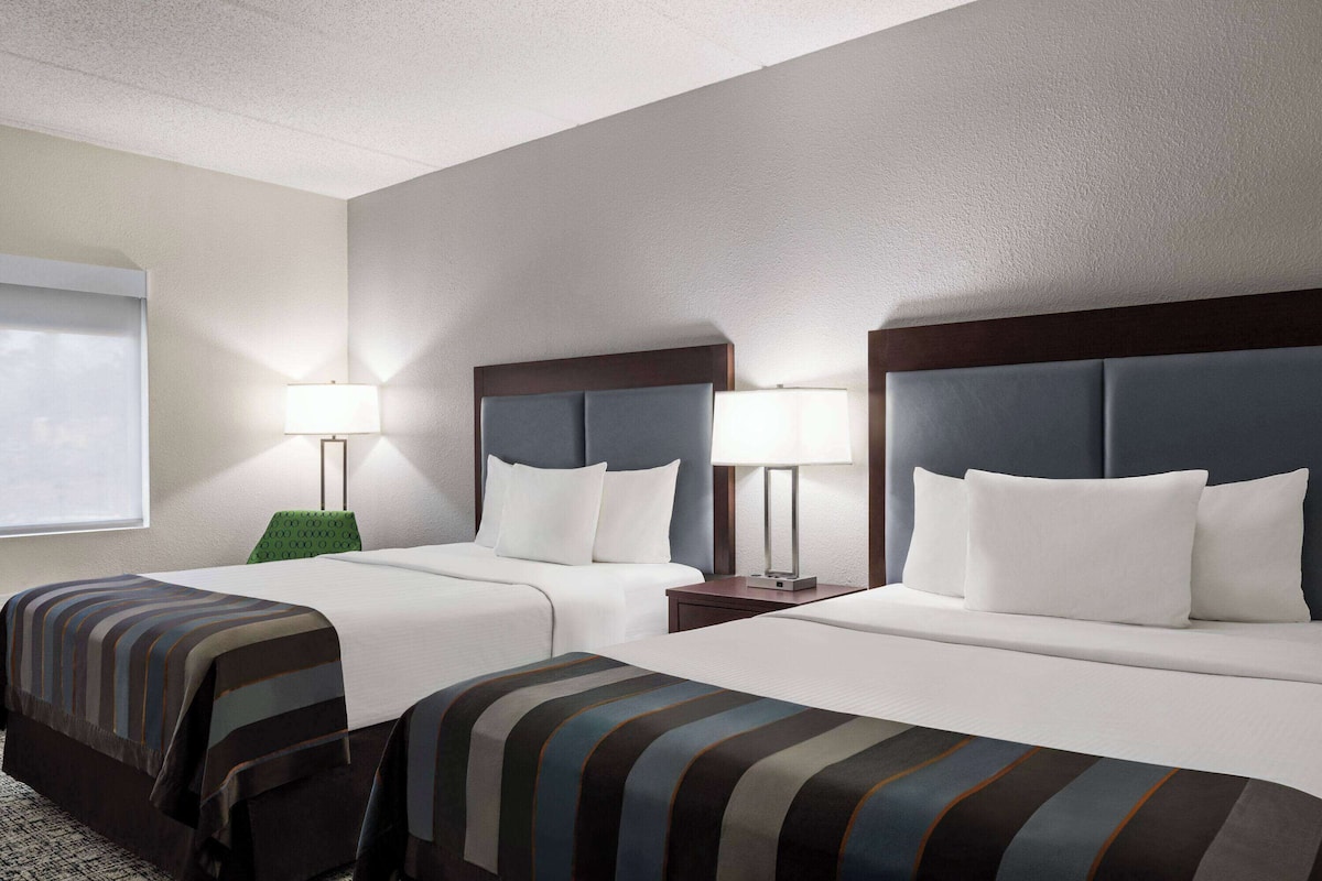 A hotel room with two beds, each with a headboard, a bedside table with a lamp, and a window with blinds.
