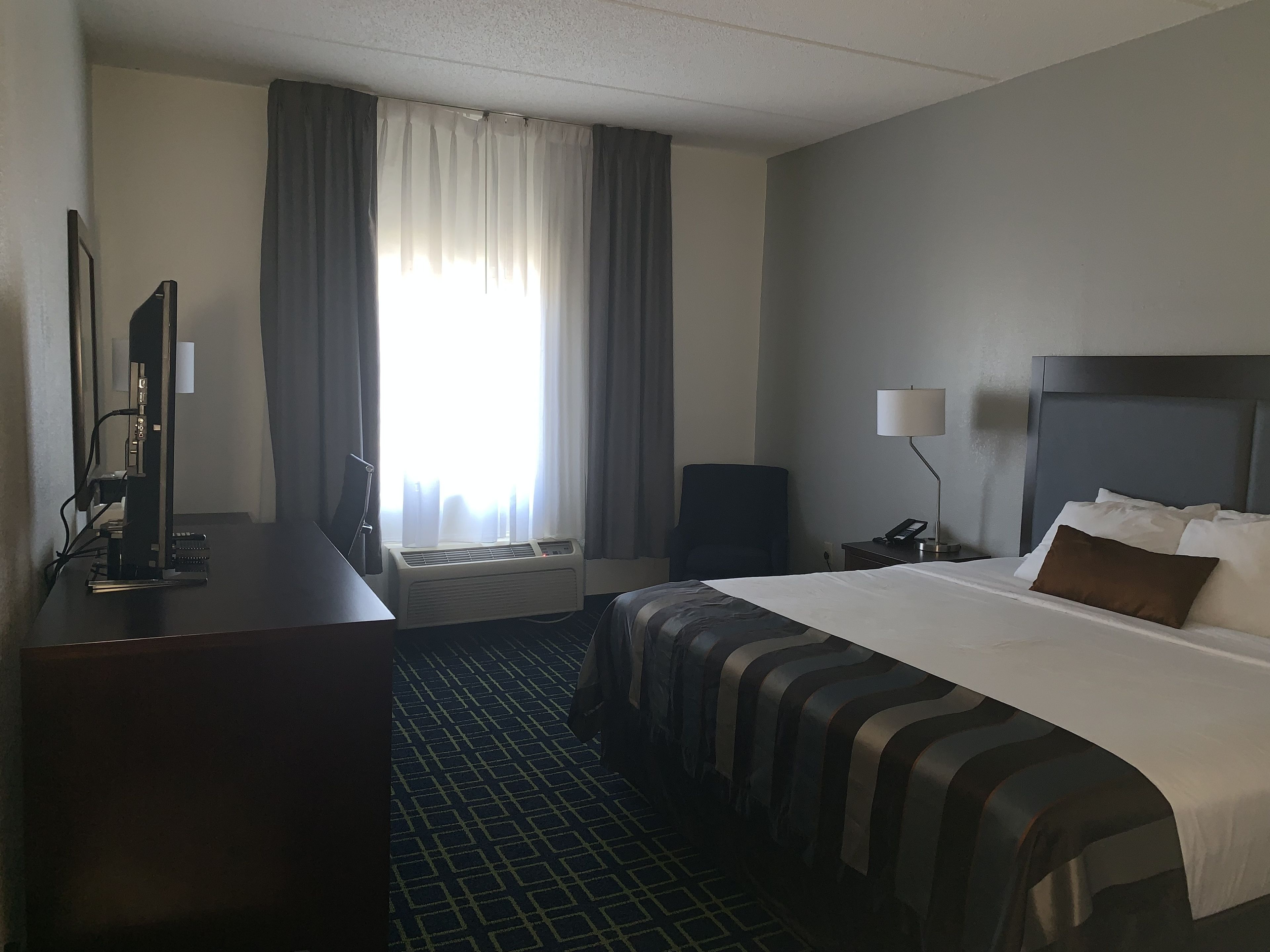 A hotel room with a bed, a desk with a computer, a chair, a bedside table with a lamp, and a window with curtains.