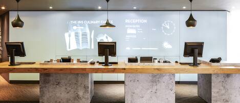 A reception area with a wooden reception desk, two computer monitors, and a projection screen displaying room rates and services.