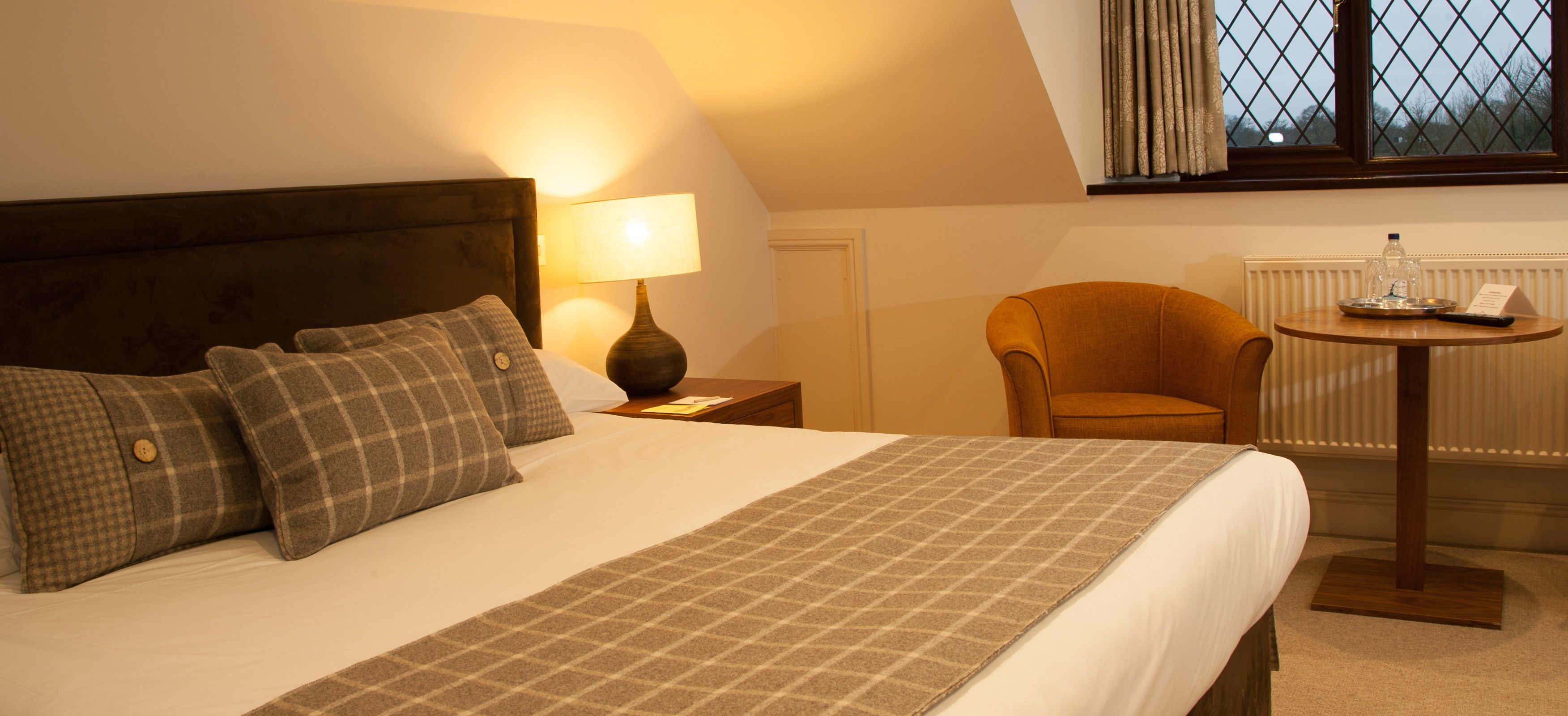A neatly made bed with a plaid blanket and pillows, a bedside lamp, a chair, and a small table with a remote and a bottle.