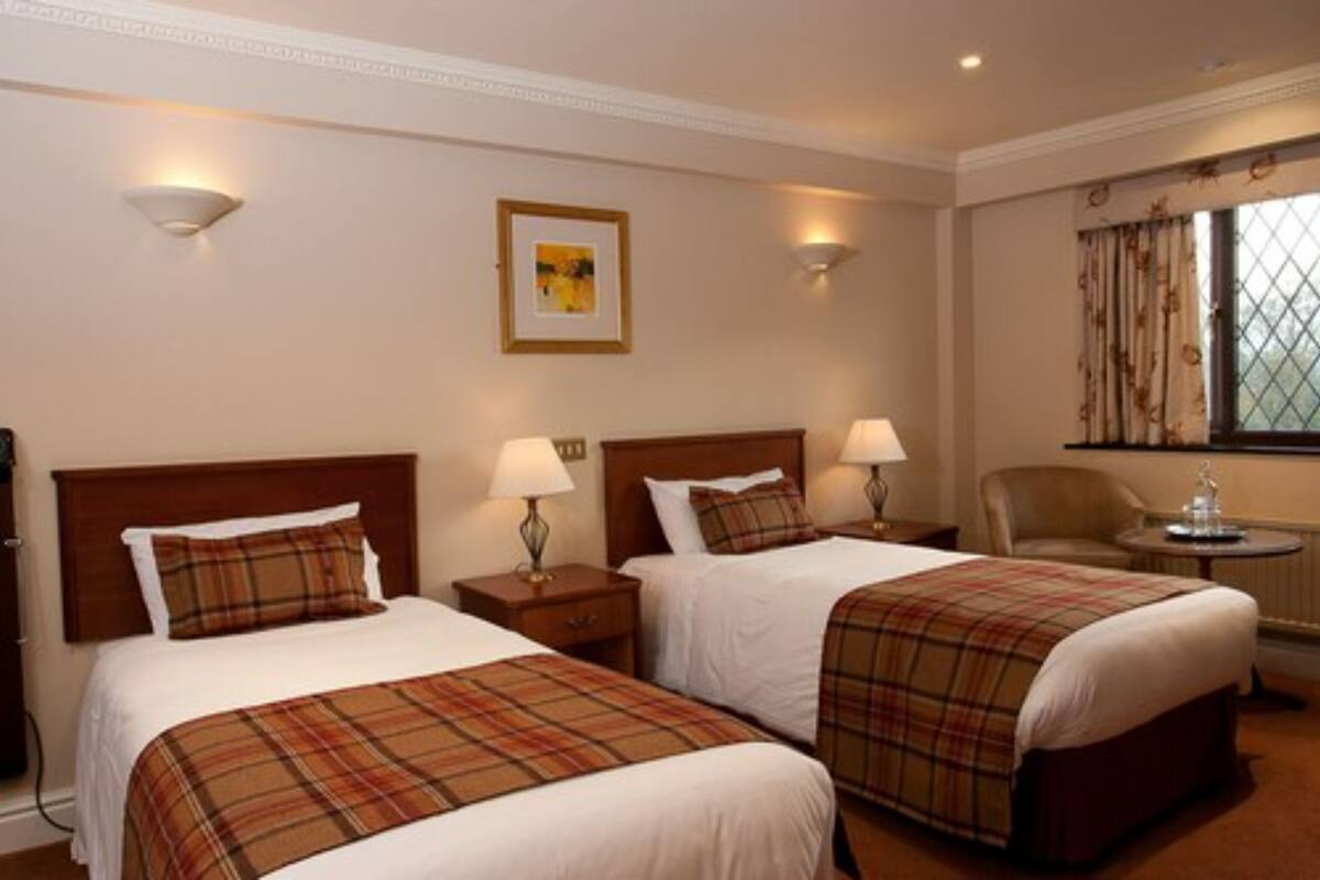 A hotel room with two beds, each with a plaid bedspread, bedside lamps, a framed picture on the wall, a window with curtains, and a small table with a lamp.