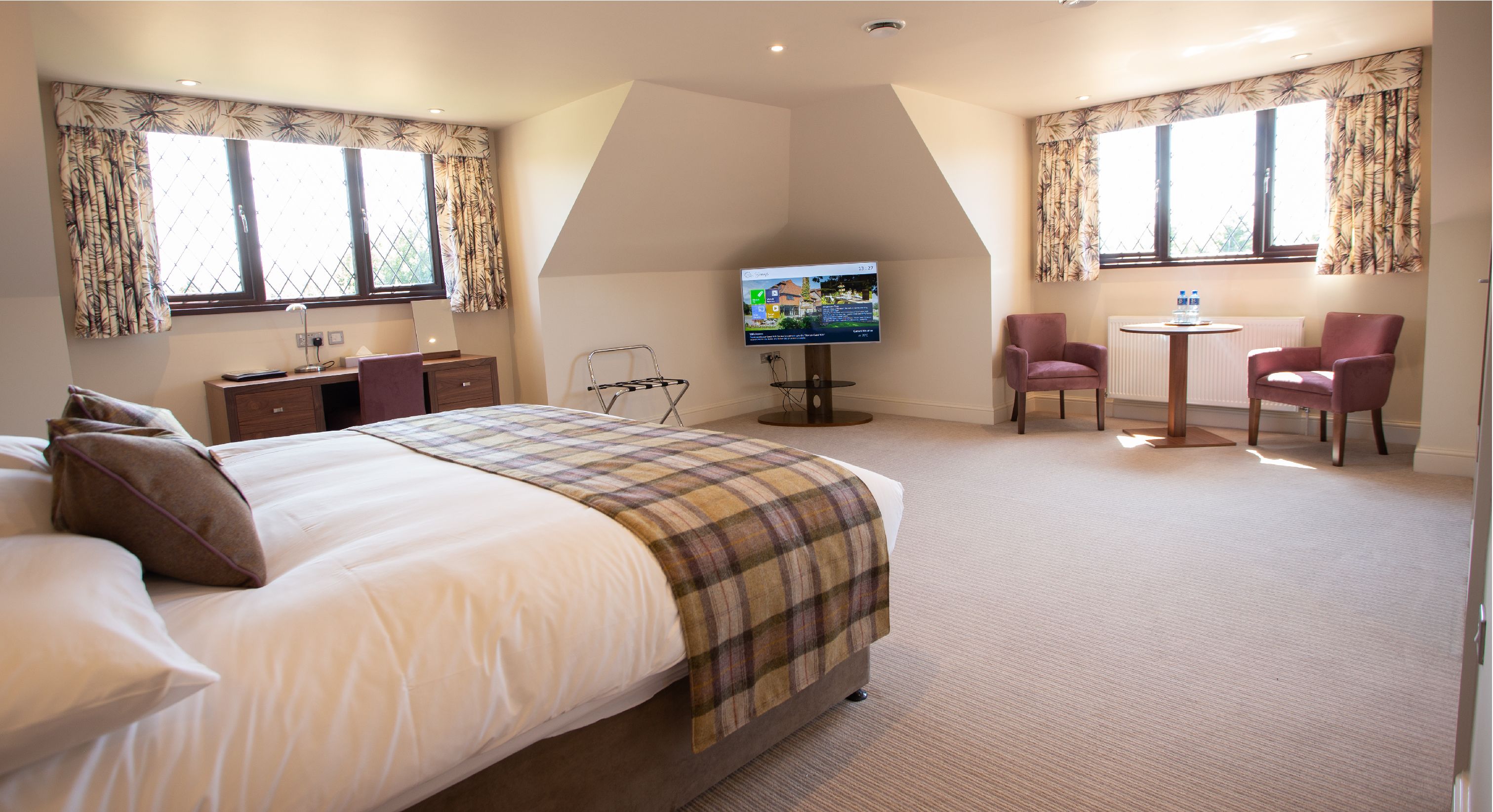 A hotel room with a large bed, a desk, a chair, a TV, and two windows with curtains.
