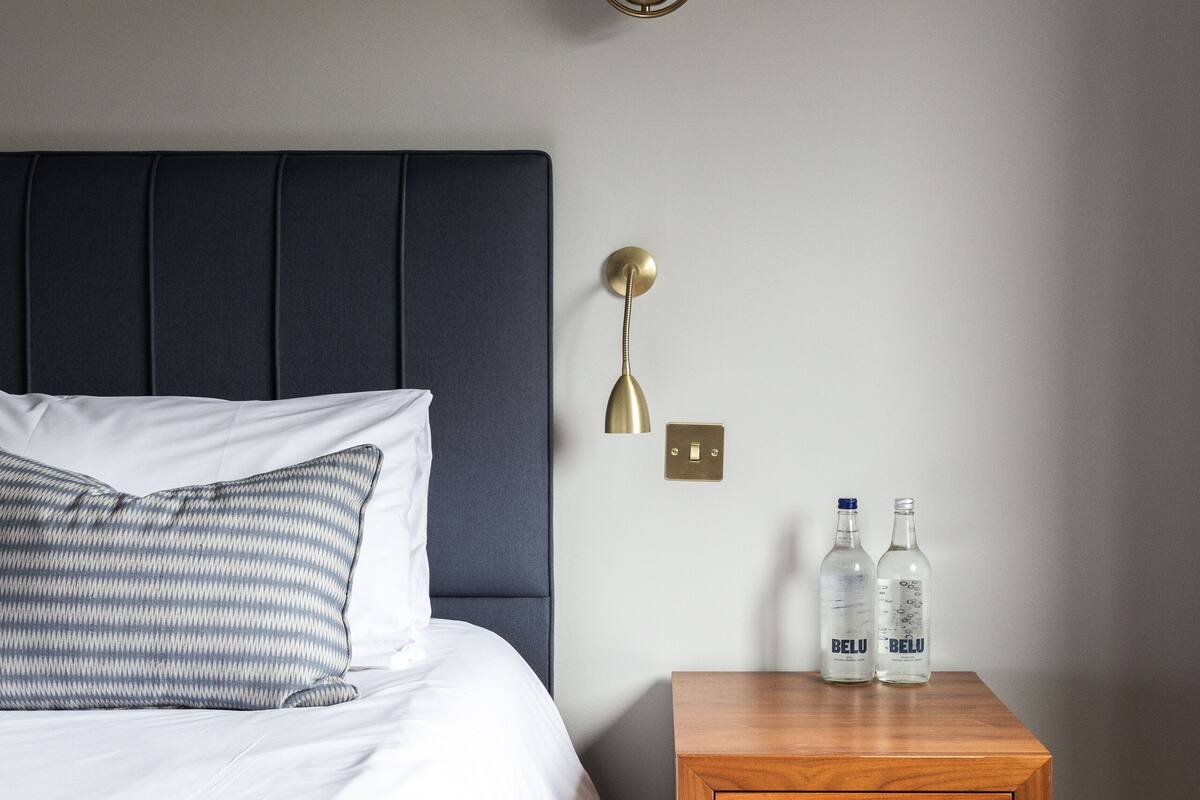 A neatly made bed with patterned pillows, a bedside table with two bottles, and a wall-mounted lamp.