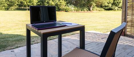 Un patio con una computadora portátil sobre una mesa, una silla y vistas a un área verde con árboles.