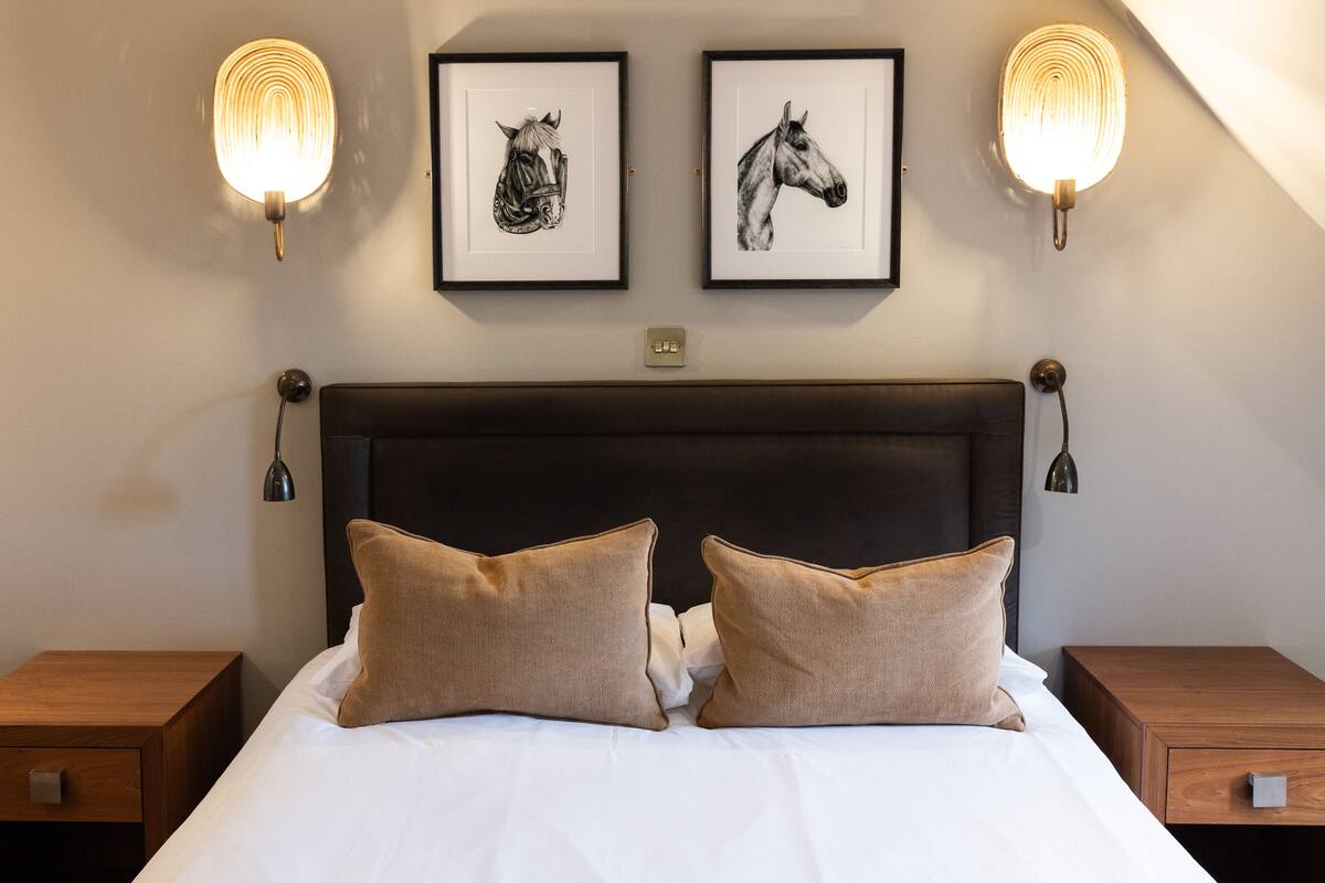 A neatly made bed with two pillows, a dark headboard, and two framed horse illustrations on the wall above.