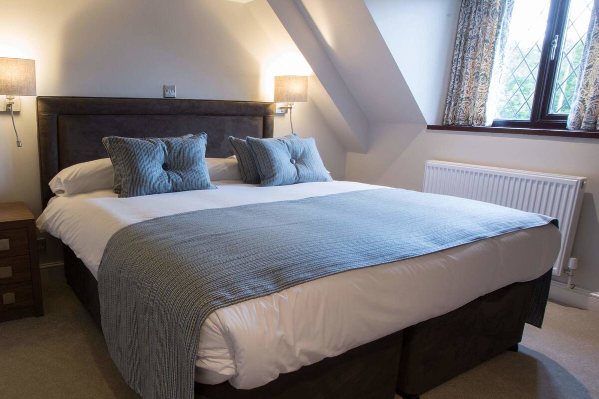 A bedroom with a large bed, two bedside lamps, a chair, and a window with curtains.
