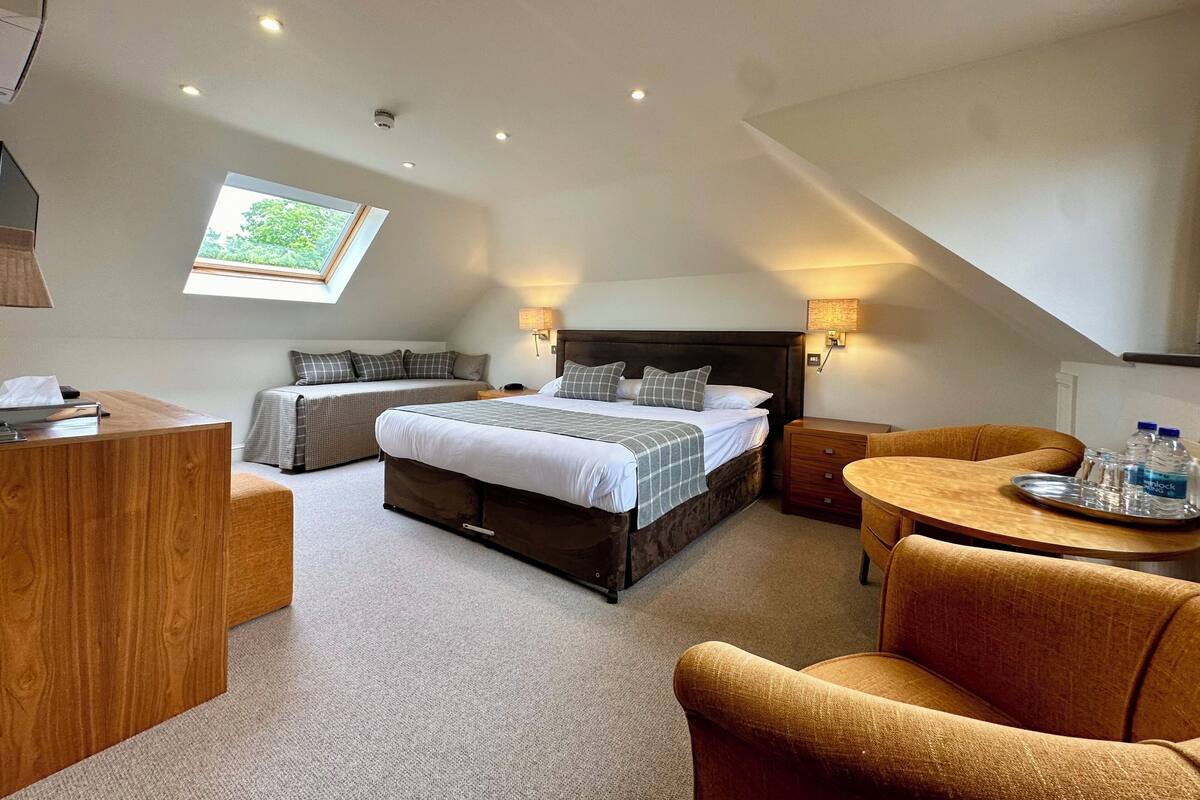 A modern hotel room with a large bed, a desk, a sofa, and a small round table with water bottles.