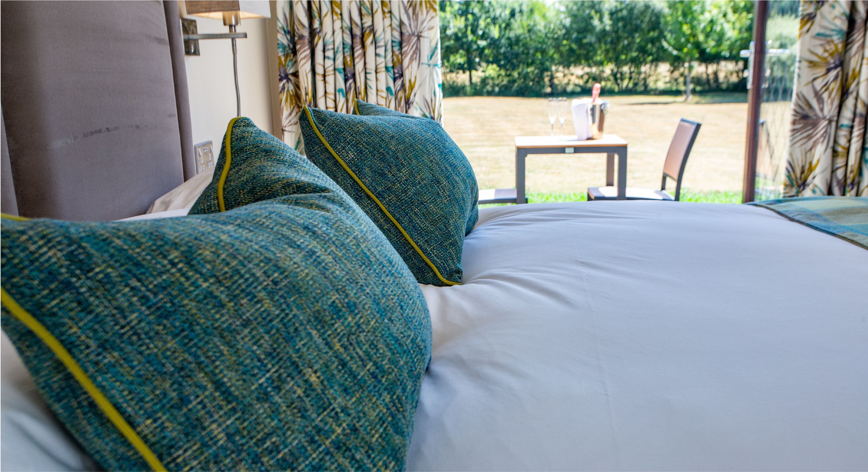 A bed with green and white bedding, a wooden nightstand, and a view of an outdoor garden area.
