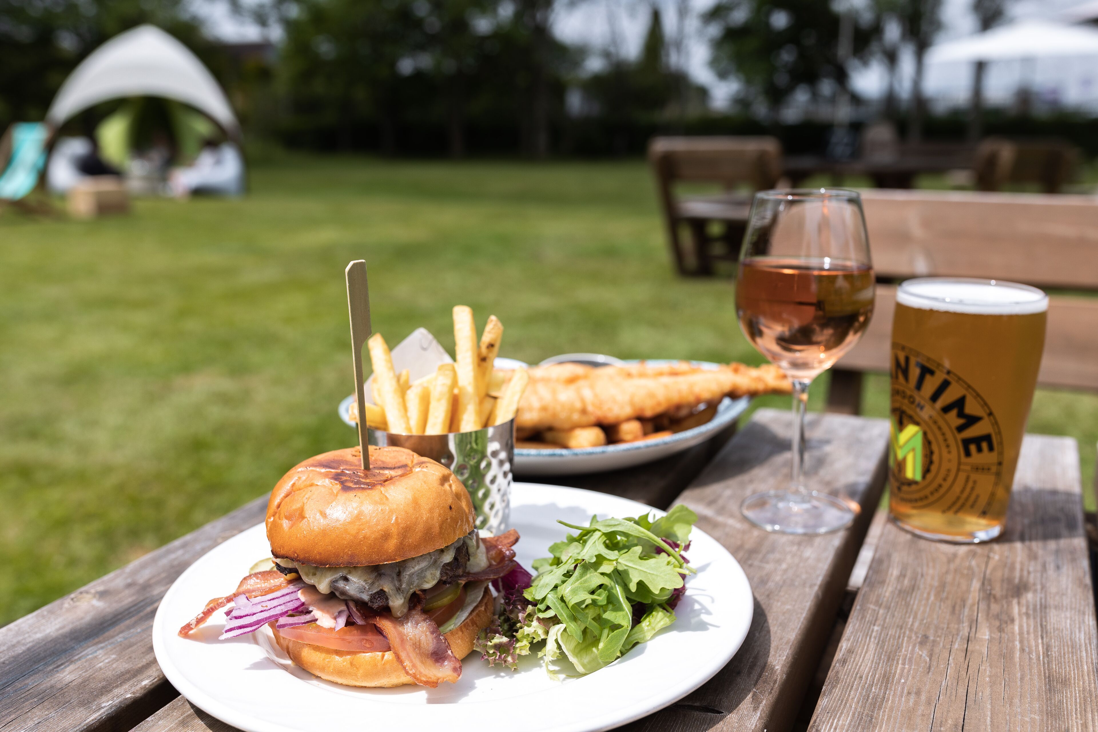 A meal consisting of a burger, fries, and a glass of beer on a wooden table in an outdoor setting.