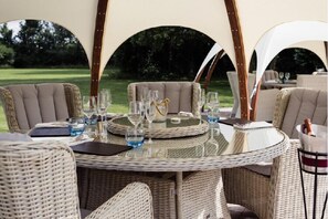 A round wicker table set for dining with chairs, a glass top, and a view of a green landscape.