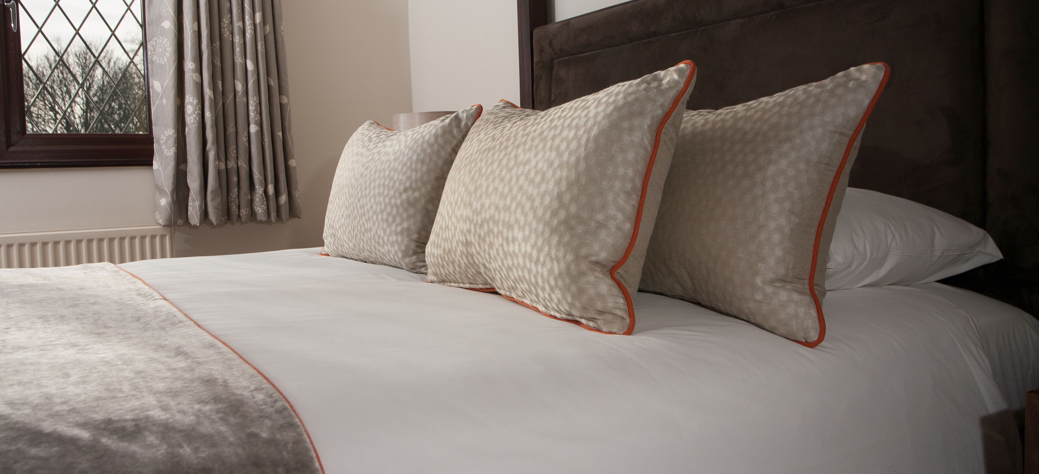 A neatly made bed with a patterned pillow and a plain pillow, a window with curtains, and a wooden headboard.