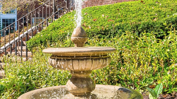 A stone fountain with water spouting from the top, surrounded by greenery and a brick building with windows in the background. - Simsbury Inn (Weatogue)