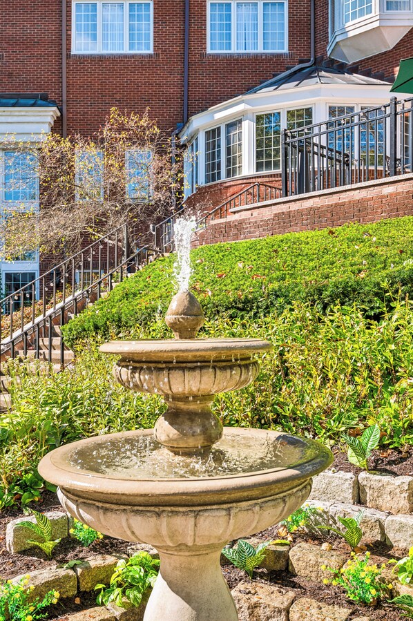 A stone fountain with water spouting from the top, surrounded by greenery and a brick building with windows in the background.