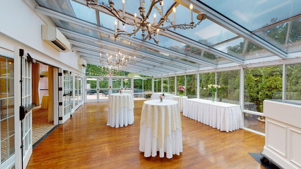 A spacious, well-lit event space with a glass ceiling, white tables, and a chandelier. - Simsbury Inn (Weatogue)