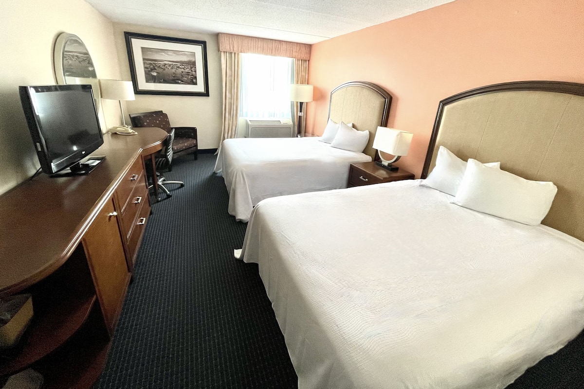 A hotel room with two beds, a desk with a television, a chair, and a window with curtains.